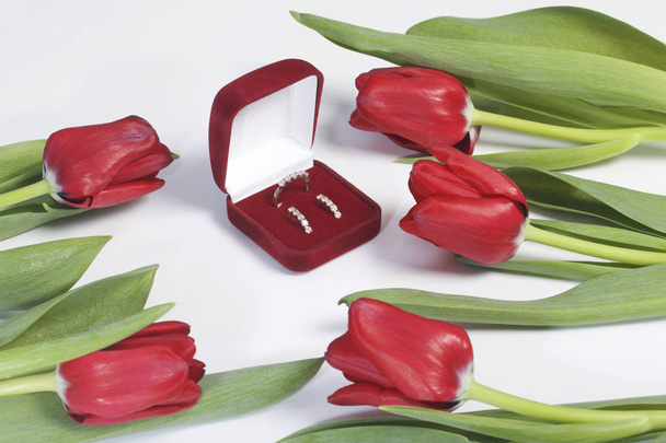 Gifts for loved ones. A bouquet of red tulips is scattered on a white surface. Nearby is a red velvet box with a ring and earrings. - Photo, image