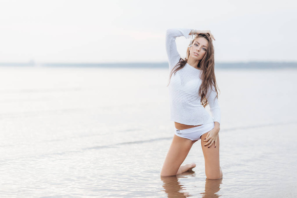 Attraente giovane modello femminile modelli al mare contro bella vista sul mare e cielo limpido, indossa bikini, si alza in ginocchio, gode di meravigliosa natura e solitudine. Perfetta forma del corpo e fitness
 - Foto, immagini