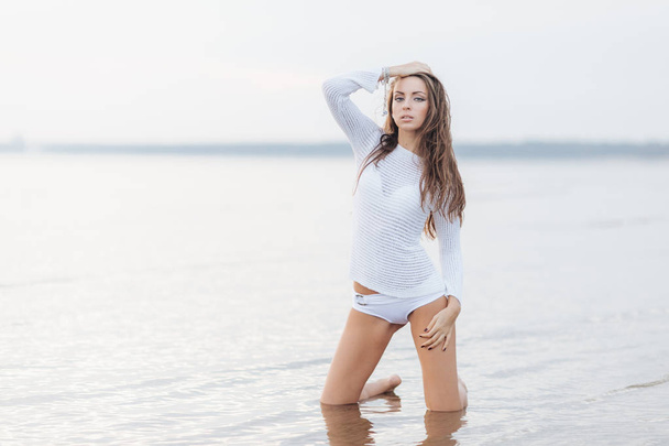 Outdooor view of good looking brunette female with make up, dressed in white bikini, poses at camera near calm sea background, has fit body shape, feels free and relaxed. Woman at seashore - Photo, Image