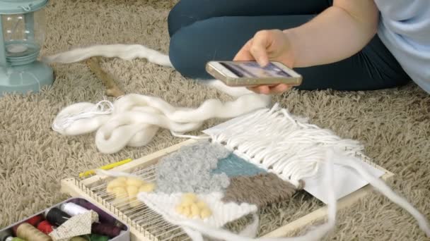Žena spřádá na tkalcovském stavu krásnou výšivkou vyrobené z nití, v domácím studiu fotografie na telefonu - Záběry, video