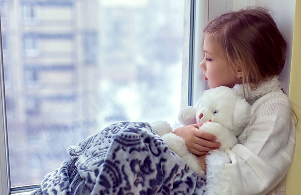 niedliches kleines Mädchen, das einen Teddybär umarmt. Ein süßes Baby im Zimmer sitzt im Winter am Fenster - Foto, Bild