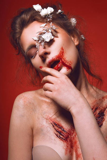 Brunette with white flowers in her hairstyle and with bloody scars on her chest on red background - Фото, зображення