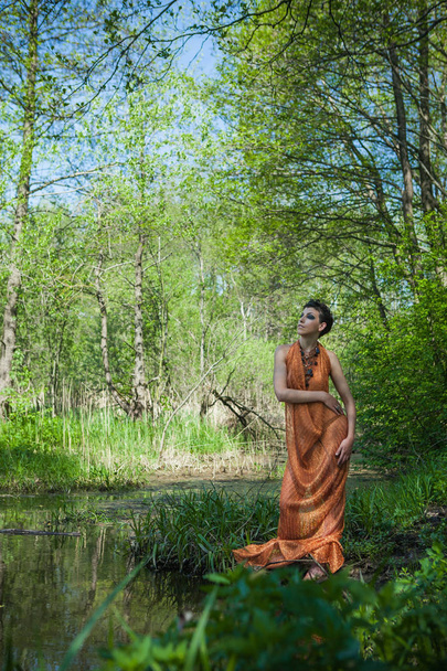 slim brunette in an orange dress is standing on the shore of a forest pond - Фото, зображення