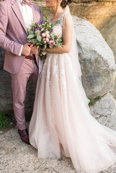 Wedding. The girl in a beige dress and a guy in a suit are holding a beautiful bouquet of white, purple, lilac flowers and greenery. Wedding rustic bouquet with lilac roses. Wedding details - Photo, Image