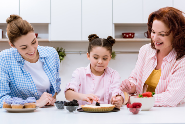 trois générations de femmes heureuses décorer dessert avec des baies ensemble à la cuisine
 - Photo, image