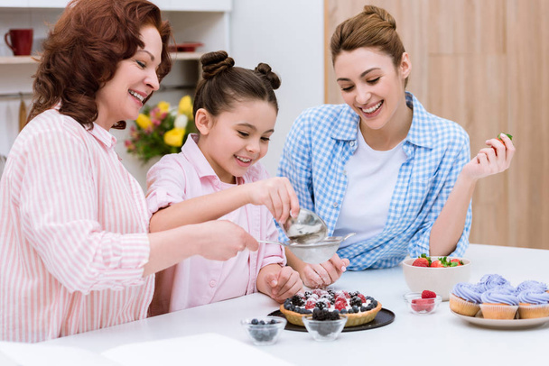 three generations of women pouring sugar onto tart with berries together at kitchen - Фото, изображение