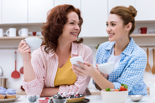 happy young woman and her mother drinking coffee with sweets and using smartphone together - Zdjęcie, obraz