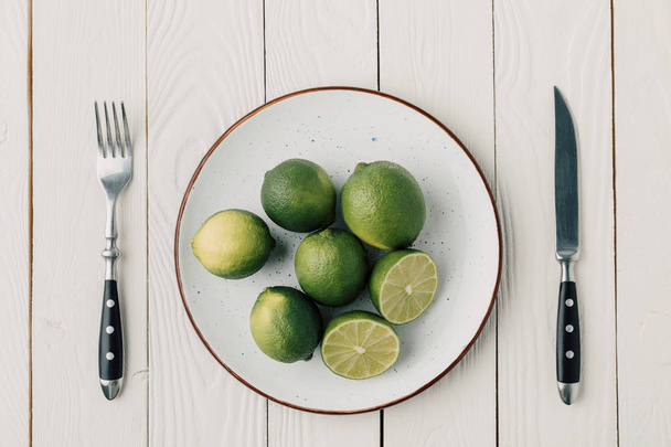 top view of green limes on plate and cutlery on white wooden background - Foto, Bild