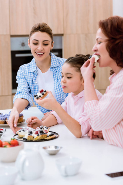 three generations of women eating delicious tart with berries at kitchen - Foto, Bild