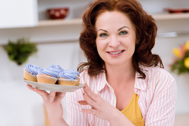 close-up portrait of beautiful mature woman with plate of blueberry cupcakes at kitchen - Fotó, kép