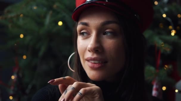 Charming model poses in red winter hat before a Christmas tree - Кадри, відео