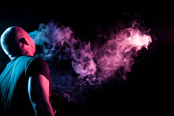 A young man exhales a cloud of colored smoke of pink and  purple color on a black isolated color.  Guy is admiting puffs of steam from the electronic cigarette.  - Foto, Imagen