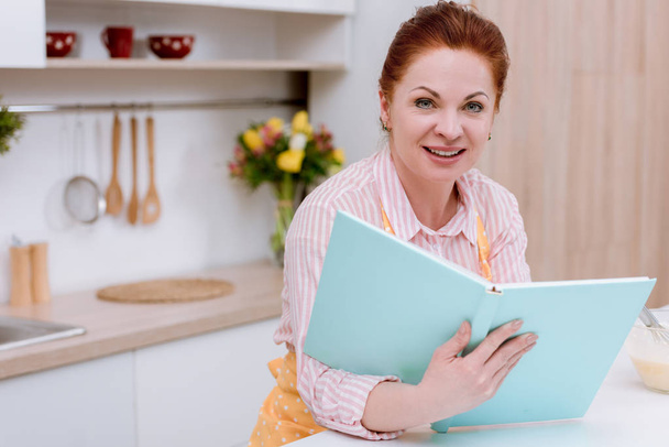 happy mature woman in apron with recipe book - 写真・画像