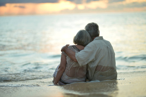 Achteraanzicht van bejaarde echtpaar staande aan zandstrand tijdens zonsondergang - Foto, afbeelding