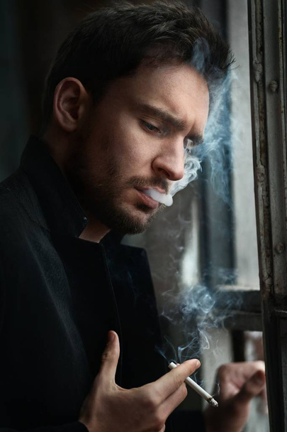 Mysterious handsome man with sneaky look. man with beard smiling. sexy guy in a dark jacket with confident face. Sexy and smart man with beautiful eyes. businessman on a dark romantic background smoking cigarette - Photo, Image