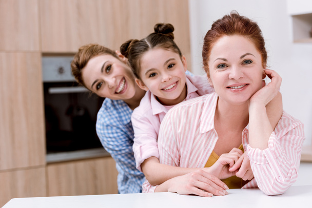 drei Generationen glücklicher schöner Frauen, die in einer Reihe in der Küche stehen und in die Kamera schauen - Foto, Bild