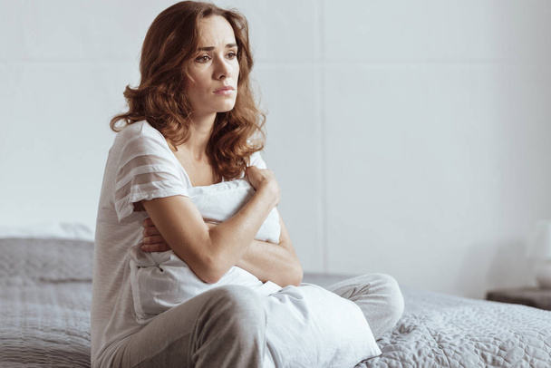Depressed mature woman embracing pillow while sitting on bed - Foto, Imagen