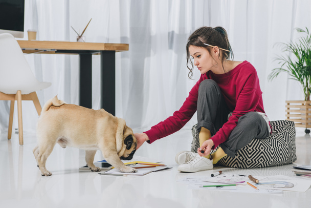 Attractive young girl cuddling pug on floor among sketches - Foto, imagen