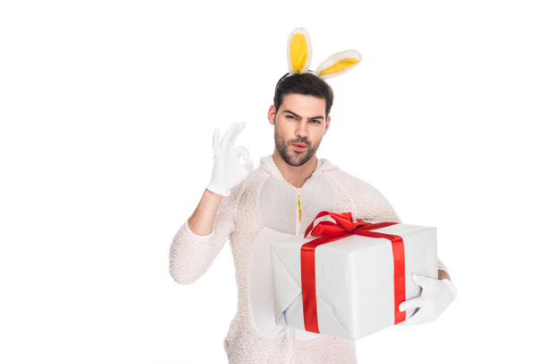 Улыбающийся человек в костюме кролика держит подарочную коробку и показывает ОК знак изолирован на белом, пасхальная концепция
 - Фото, изображение