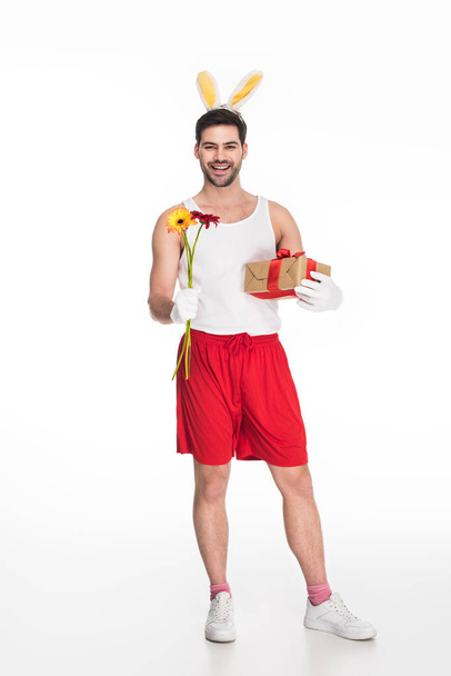 Smiling man with bunny ears holding flowers and gift box isolated on white, easter concept - Fotografie, Obrázek