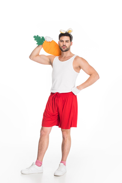 View of man with bunny ears holding big carrot on shoulder isolated on white, easter concept - Photo, Image