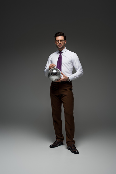 Young businessman in glasses holding glass disco ball on grey - Fotó, kép