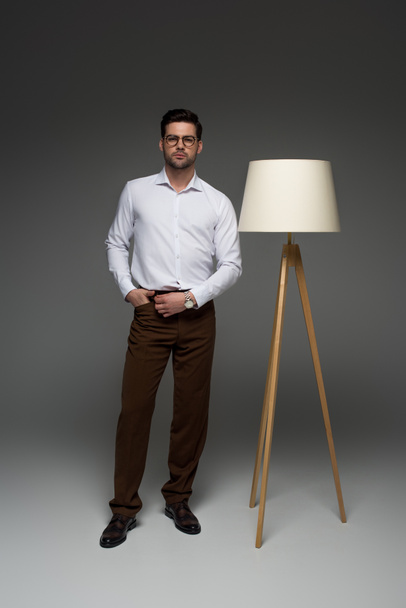 Vista frontal del hombre de negocios de pie con lámpara de pie al lado en gris
 - Foto, imagen