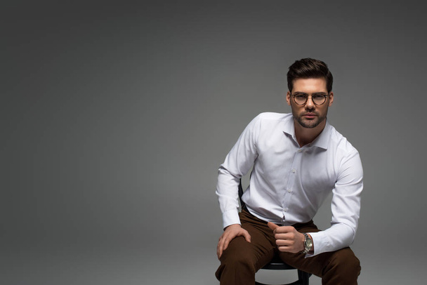 stylish businessman in glasses sitting on chair and looking at camera isolated on grey - Foto, Imagem
