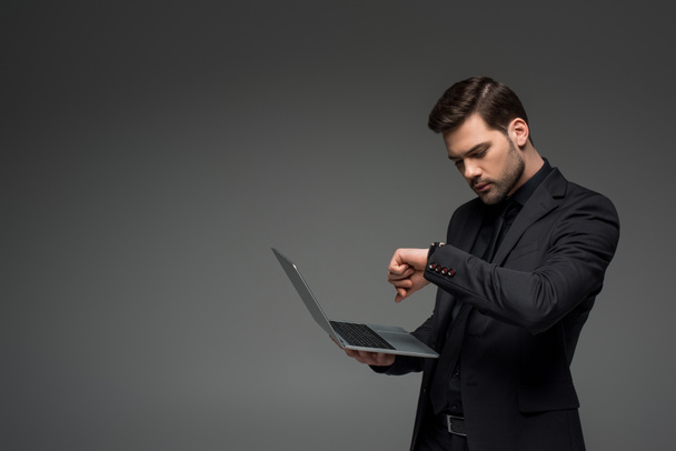 Liikemies katselee rannekelloa kannettava tietokone kädessä eristettynä harmaalla
 - Valokuva, kuva