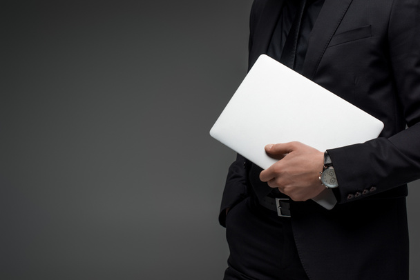 Partial view of businessman holding laptop in hand isolated on grey - Foto, imagen