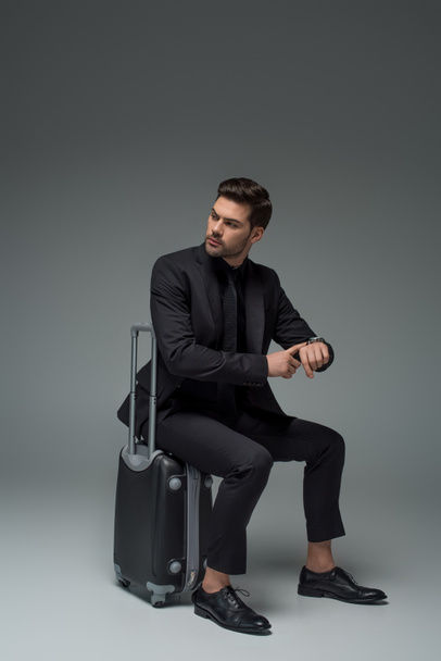 Business tourist sitting on suitcase and pointing at wristwatch on grey - Fotografie, Obrázek