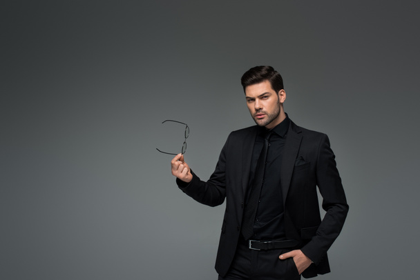 Young stylish man in formal suit holding glasses isolated on grey - Photo, image