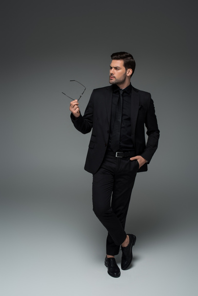 Stylish man in black suit posing with glasses in hand on grey - 写真・画像