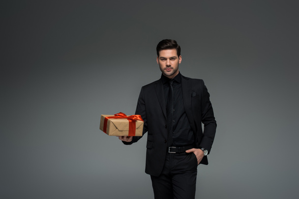 Stylish man in black suit holding gift box isolated on grey, international womens day concept - Foto, Imagen