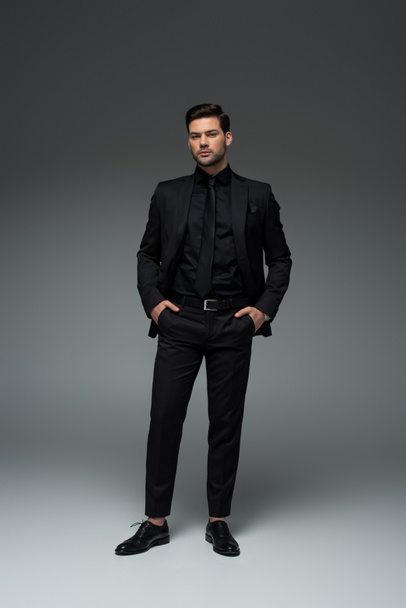 Hombre de elegante traje negro de pie con los dedos en los bolsillos en gris
 - Foto, Imagen