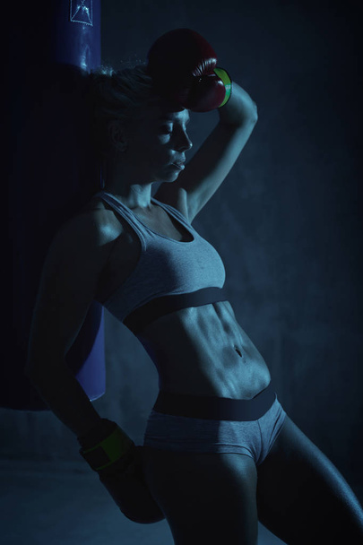 Blue lighted portrait of sexy hot blonde boxer woman in red boxing gloves and grey sports bra and panties standing near blue punching bag with hand on forehead, tired - Foto, Imagen
