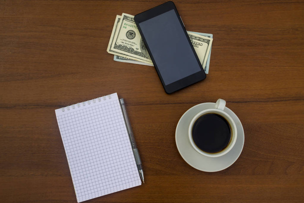 Cup of coffee, smartphone, dollars, notepad and pen on wooden desk - Foto, Imagem