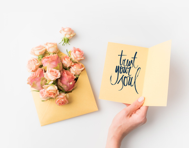 hand holding paper with TRUST YOUR SOUL sign beside pink flowers in envelope isolated on white - Фото, изображение