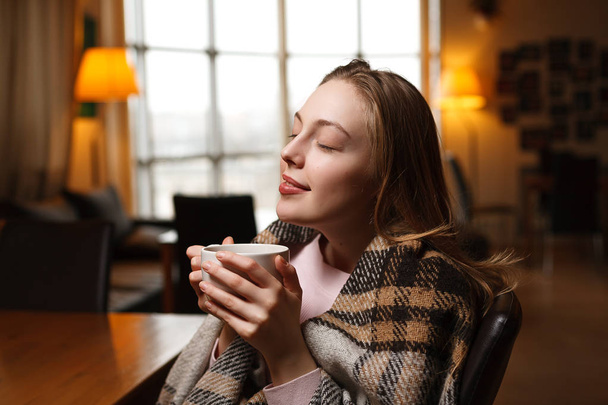 Prachtige gelukkige vrouw drinkt koffie in een café achtergrond. Overdekte gezellige plaid. Achtergrond wazig café. - Foto, afbeelding