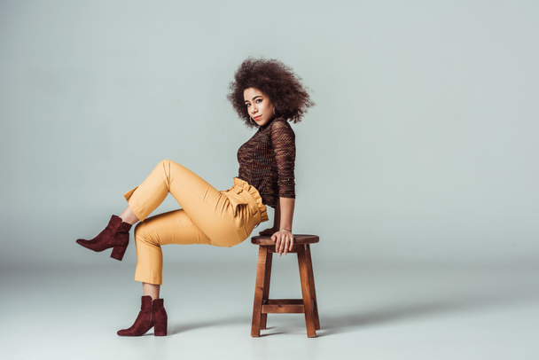 side view of african american woman in retro clothes leaning on chair on grey - Фото, изображение