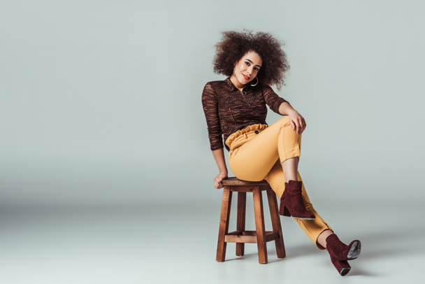 smiling african american woman in retro clothes posing on chair on grey - Foto, immagini