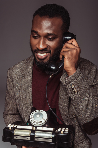 smiling african american retro styled man talking by vintage stationary telephone isolated on grey - Photo, Image