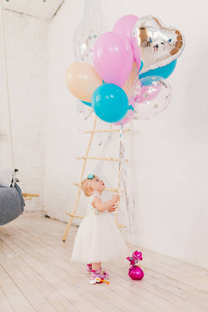 白い豪華なドレスでの小さな女の赤ちゃんは装飾の階段の近くの家のリビング ルームで立っているし、鮮やかな色の風船の束を伸ばします。子供の誕生日パーティーのコンセプト - 写真・画像