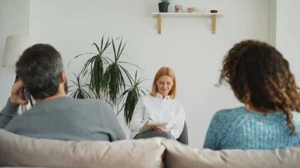 Female professional psychologist listening and writing notes in notebook while young woman arguing about boyfriend talking by phone during visit psychotherapy office indoors - Filmati, video