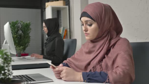 Young beautiful girl in pink hijab sits in office and uses smartphone. Girl in black hijab in the background. Arab women in the office. 60 fps - Кадри, відео