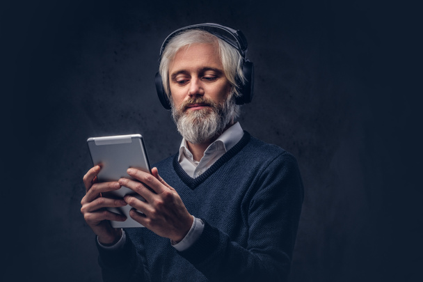 Studio portrait of a handsome senior man using a tablet with headphones over a dark background. - 写真・画像