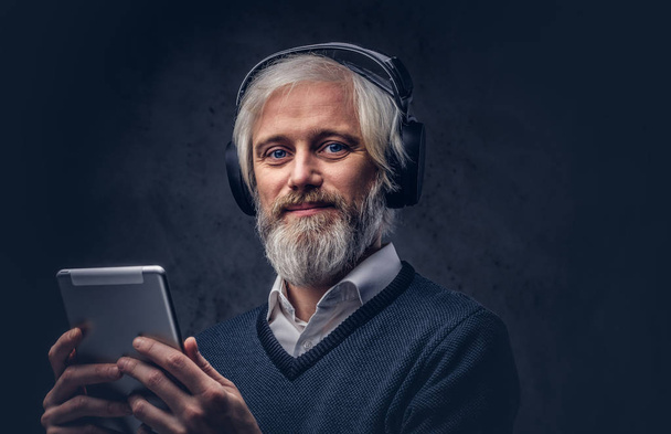 Retrato de estudio de un hombre mayor guapo usando una tableta con auriculares sobre un fondo oscuro
. - Foto, imagen