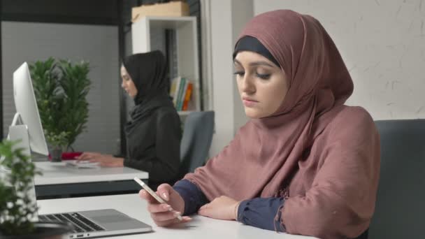 Young beautiful girl in pink hijab sits in office and uses smartphone. Girl in black hijab in the background. Arab women in the office. 60 fps - Video