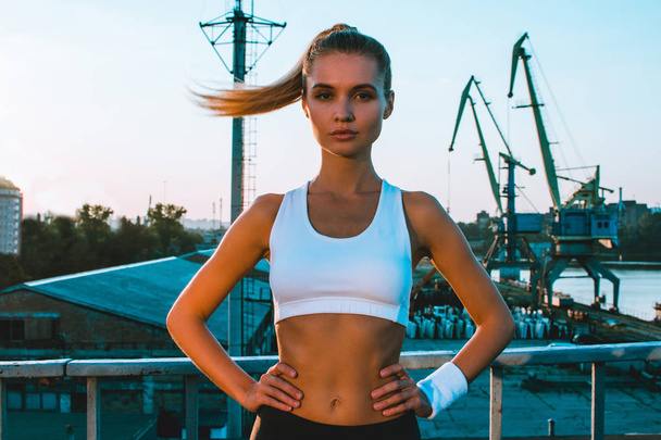 Morning run. Young beautiful woman in sportswear looking at camera against industrial city view - Photo, Image