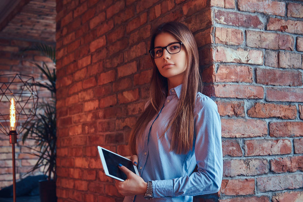 Portrait of an attractive charming businesswoman in glasses and blue shirt holds a tablet leaning against on a brick wall in a room with loft design. - Photo, Image